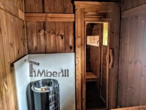TimberIN Nordic Oval™ Terrace – interior: estufa de sauna delante de un panel de protección térmica, puerta y banco de madera