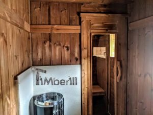 TimberIN Nordic Oval™ Terrace – interior: estufa de sauna delante de un panel de protección térmica, puerta y banco de madera