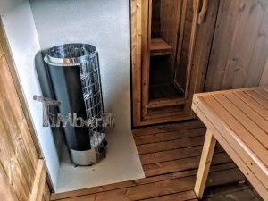 TimberIN Nordic Oval™ Terrace – interior: estufa de sauna delante de un panel de protección térmica, puerta y banco de madera