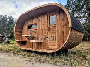 TimberIN Nordic Oval™ Terrace – sauna de exterior, vista de tres cuartos: cubierta de tejas negras, fachada de madera termotratada y porche cubierto con asientos abatibles, molino de viento al fondo