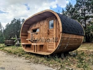 TimberIN Nordic Oval™ Terrace – sauna de exterior, vista de tres cuartos: cubierta de tejas negras, fachada de madera termotratada y porche cubierto con asientos abatibles, molino de viento al fondo