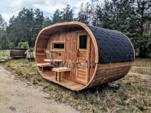 TimberIN Nordic Oval™ Terrace – sauna de exterior, vista de tres cuartos: cubierta de tejas negras, fachada de madera termotratada y porche cubierto con asientos abatibles, molino de viento al fondo