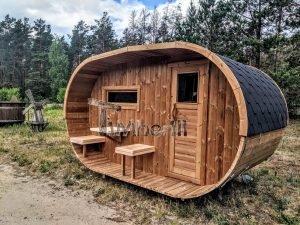TimberIN Nordic Oval™ Terrace – sauna de exterior, vista de tres cuartos: cubierta de tejas negras, fachada de madera termotratada y porche cubierto con asientos abatibles, molino de viento al fondo