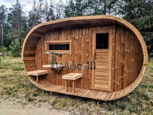 TimberIN Nordic Oval™ Terrace – sauna de exterior, vista frontal: fachada de madera termotratada con puerta acristalada, ventana lateral y porche cubierto con asientos abatibles y mesita, pinos al fondo