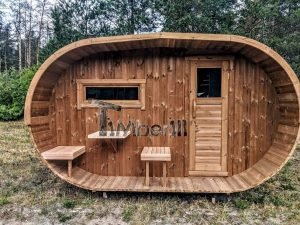 TimberIN Nordic Oval™ Terrace – sauna de exterior, vista frontal: fachada de madera termotratada con puerta acristalada, ventana lateral y porche cubierto con asientos abatibles y mesita, pinos al fondo