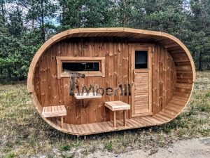 TimberIN Nordic Oval™ Terrace – sauna de exterior, vista frontal: fachada de madera termotratada con puerta acristalada, ventana lateral y porche cubierto con asientos abatibles y mesita, pinos al fondo