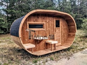 TimberIN Nordic Oval™ Terrace – sauna de exterior, vista de tres cuartos: cubierta de tejas negras, fachada de madera termotratada y porche cubierto con asientos abatibles y mesita