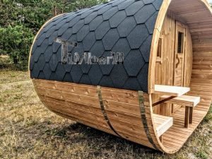 TimberIN Nordic Oval™ Terrace – sauna de exterior, vista trasera de tres cuartos: cubierta de tejas negras sobre la estructura curva de madera termotratada, asientos del porche visibles delante
