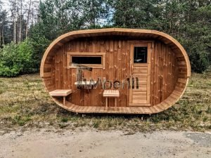 TimberIN Nordic Oval™ Terrace – sauna de exterior, vista frontal: fachada de madera termotratada con puerta acristalada, ventana lateral y porche cubierto con asientos abatibles y mesita, pinos al fondo