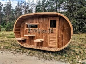 TimberIN Nordic Oval™ Terrace – sauna de exterior, vista frontal: fachada de madera termotratada con puerta acristalada, ventana lateral y porche cubierto con asientos abatibles y mesita, pinos al fondo