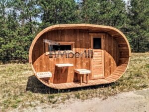 TimberIN Nordic Oval™ Terrace – sauna de exterior, vista frontal: fachada de madera termotratada con puerta acristalada, ventana lateral y porche cubierto con asientos abatibles y mesita, pinos al fondo