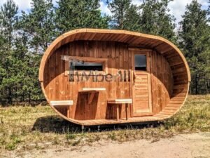 TimberIN Nordic Oval™ Terrace – sauna de exterior, vista frontal: fachada de madera termotratada con puerta acristalada, ventana lateral y porche cubierto con asientos abatibles y mesita, pinos al fondo