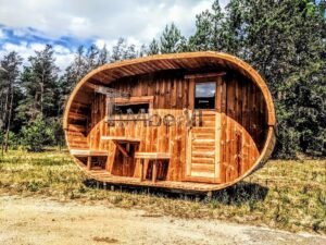 TimberIN Nordic Oval™ Terrace – sauna de exterior, vista frontal: fachada de madera termotratada con puerta acristalada, ventana lateral y porche cubierto con asientos abatibles y mesita, pinos al fondo