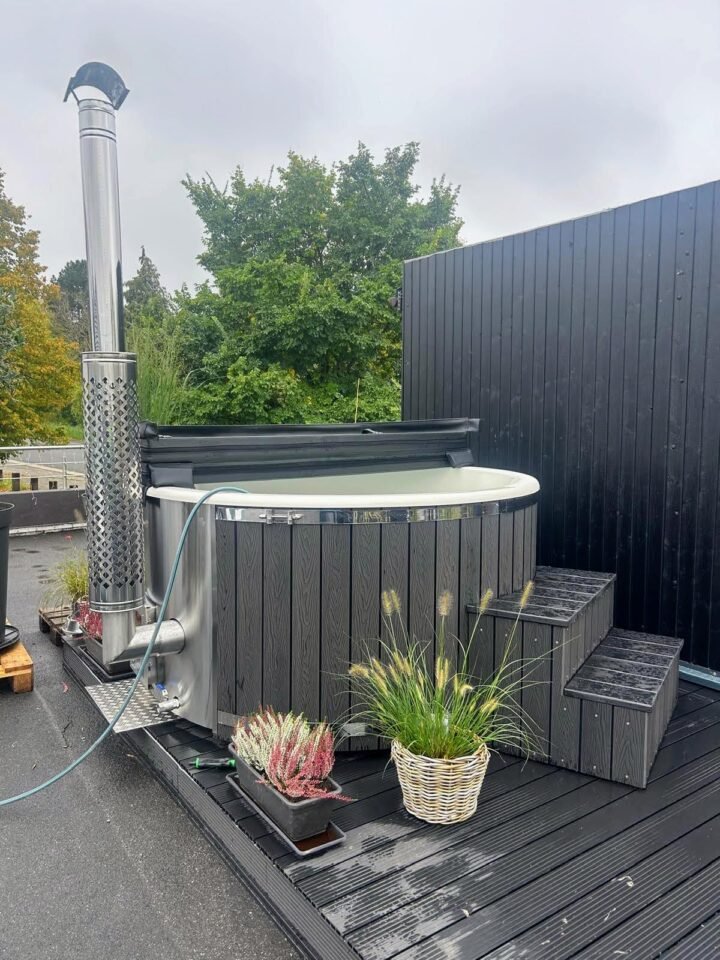 Jacuzzi gris con cubeta clara y estufa de leña integrada de acero inoxidable, con macetas de flores en una terraza