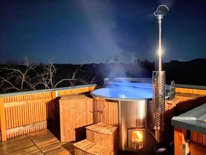 Jacuzzi exterior de madera con estufa de leña y chimenea sobre una terraza de madera al anochecer