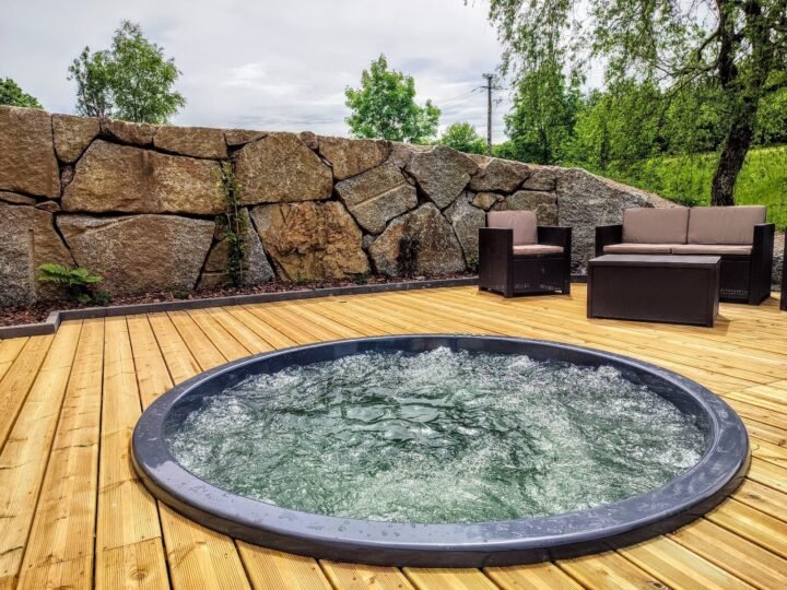 Jacuzzi redondo empotrado a ras en una gran tarima de madera, con sofás de exterior y pared de roca