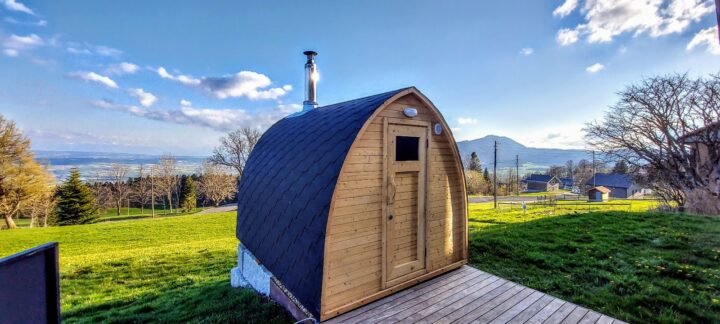 Sauna iglú de madera con tejado oscuro de tejas y chimenea de la estufa de leña, en un prado frente a las montañas