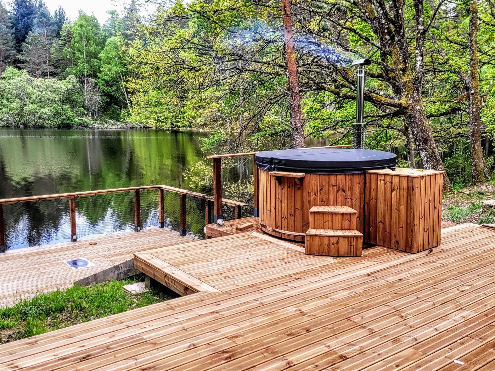 Jacuzzi redondo de madera con tapa y escalera en una terraza de madera junto a un lago en el bosque